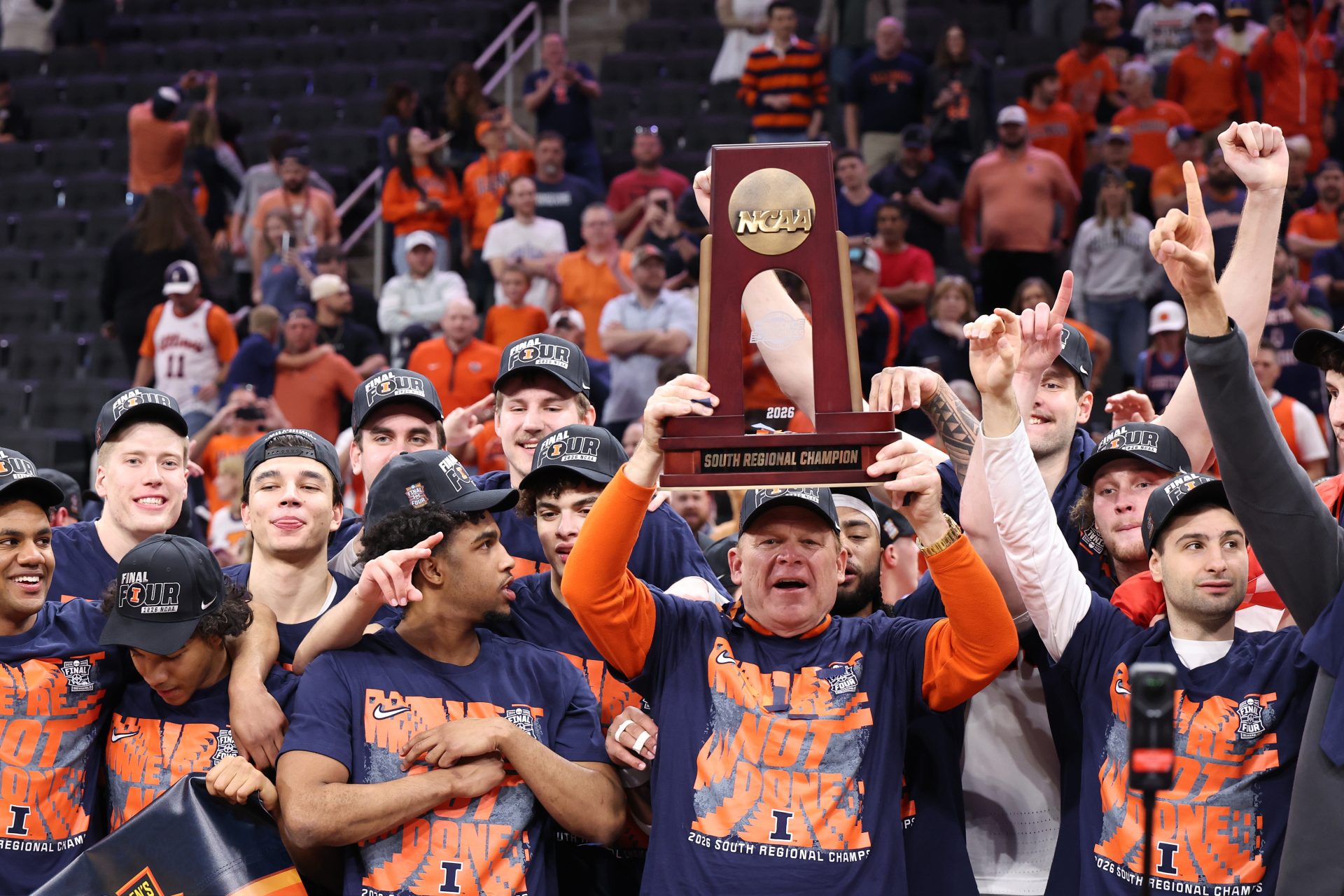Illinois Fighting Illini head coach Brad Underwood and players celebrate with the trophy on the podium after defeating the Iowa Hawkeyes in an Elite Eight game of the South Regional of the men's 2026 NCAA Tournament at Toyota Center. Source: Troy Taormina-Imagn Images